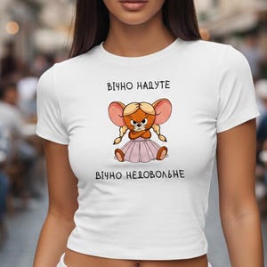Puede incluir: Top corto blanco con una ilustración de ratón de dibujos animados y el texto "ВІЧНО НАДУТЕ ВІЧНО НЕДОВОЛЬНЕ". El ratón tiene orejas rosas y una falda rosa. La modelo lleva un pantalón deportivo blanco.