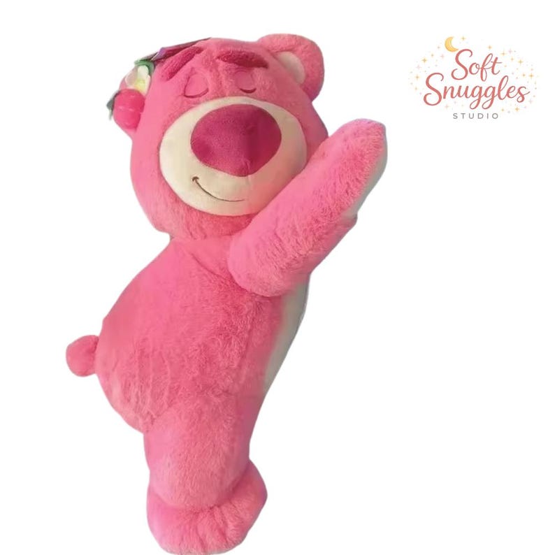 May include: A large, pink plush bear with a smiling expression and closed eyes. The bear has a white belly and a floral headpiece. The bear is holding up one arm. The text "Soft Snuggles Studio" is in the upper right corner.
