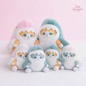 May include: A collection of plush cat toys dressed as sharks. The toys have white bodies, orange and white striped paws, and are wearing shark costumes in various colors, including blue, pink, and green. The toys have embroidered facial features and are arranged on a pink background.