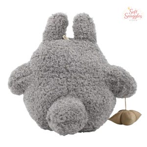 May include: A gray plush toy in the shape of a cartoon character, with large ears and a round body. The toy has a fluffy texture and a small, beige-colored accessory attached to its rear. The Soft Snuggles Studio logo is visible in the upper right corner.