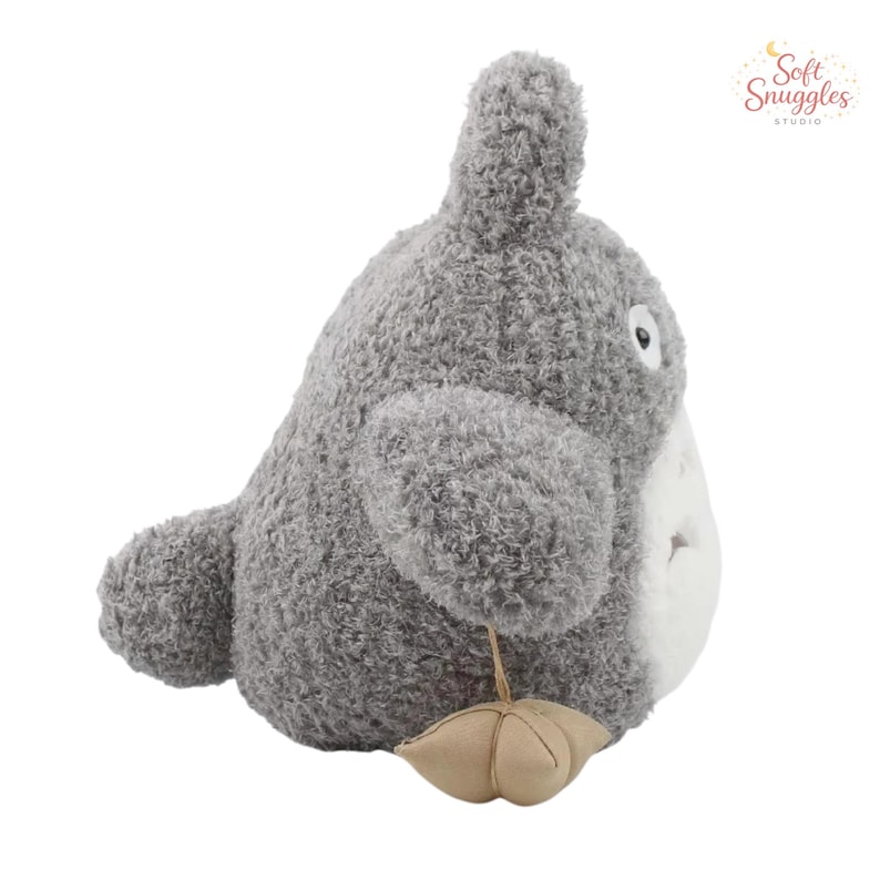 May include: A plush gray character with a white belly, large ears, and a small tan accessory. The stuffed animal has a soft, textured appearance and a friendly expression. The product is from Soft Snuggles Studio.