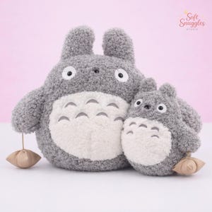 Puede incluir: Dos peluches grises de Totoro, uno grande y otro pequeño, con vientres blancos y ojos negros. Cada uno tiene una pequeña bolsa marrón colgando de su pata. El fondo es de color rosa suave.