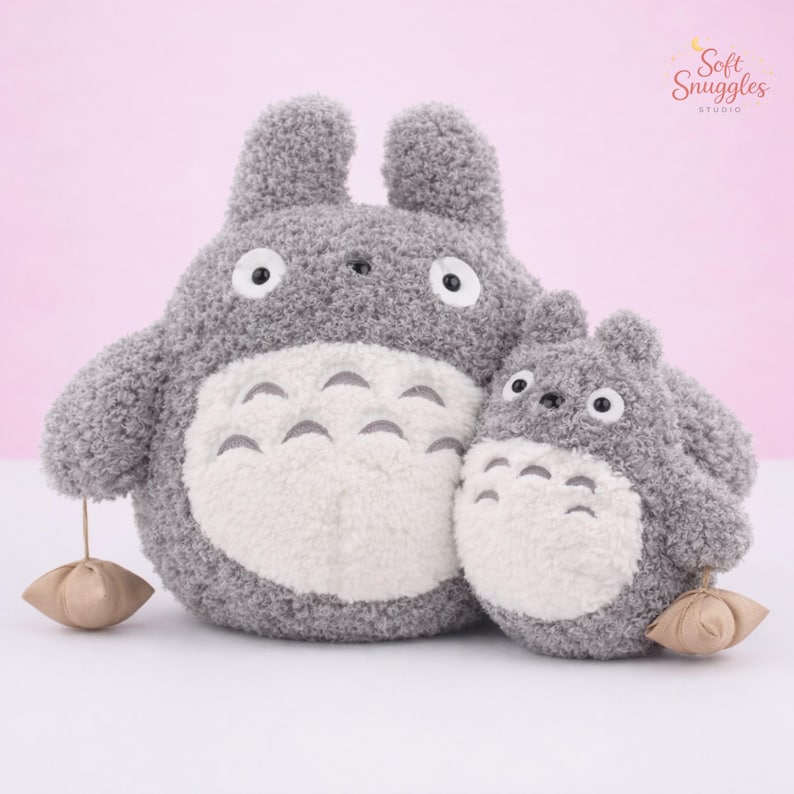 May include: Two plush gray Totoro stuffed animals, one large and one small, with white bellies and black eyes. Each has a small brown pouch hanging from its paw. The background is a soft pink.