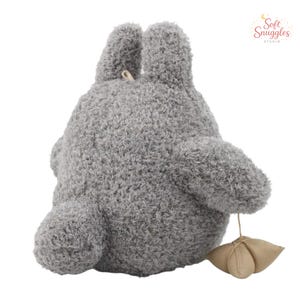 May include: A gray plush toy shaped like a cartoon character, with large ears and a rounded body. The toy has a textured, fuzzy appearance and a small, beige leaf-shaped detail attached by a string. The Soft Snuggles Studio logo is in the upper right corner.