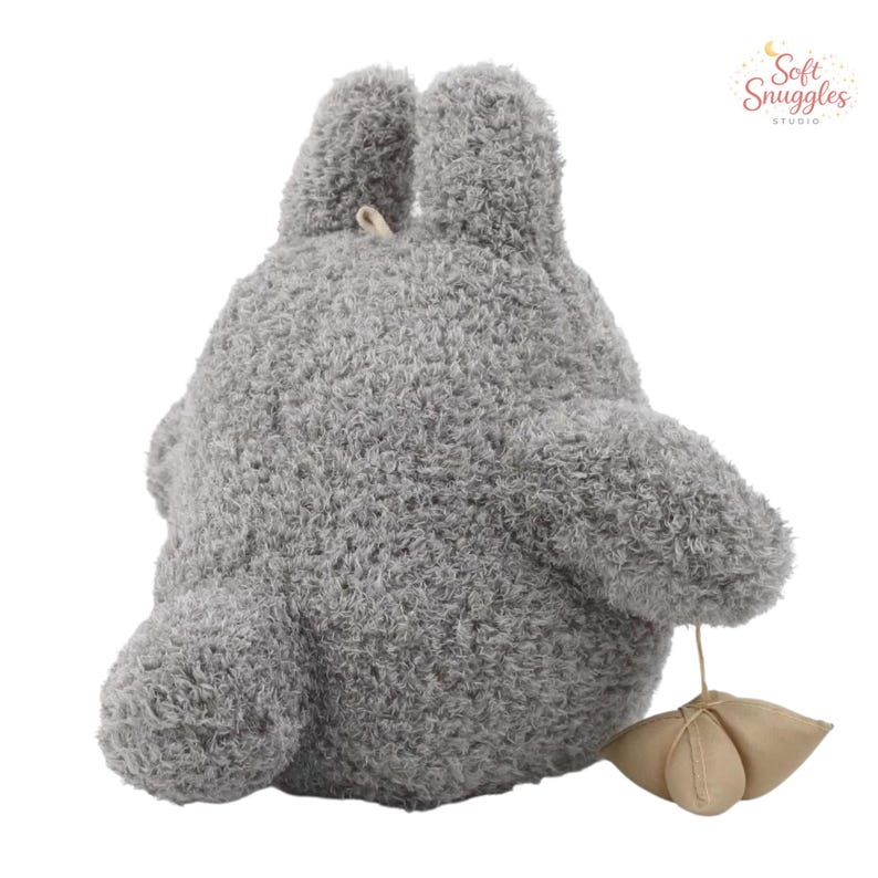 May include: A gray plush toy shaped like a cartoon character, with large ears and a rounded body. The toy has a textured, fuzzy appearance and a small, beige leaf-shaped detail attached by a string. The Soft Snuggles Studio logo is in the upper right corner.