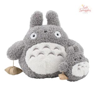 May include: A plush gray Totoro stuffed animal with a white belly and large eyes. A smaller Totoro plush is next to the larger one. The plush toys have a soft, textured appearance. The image includes the text "Soft Snuggles Studio".