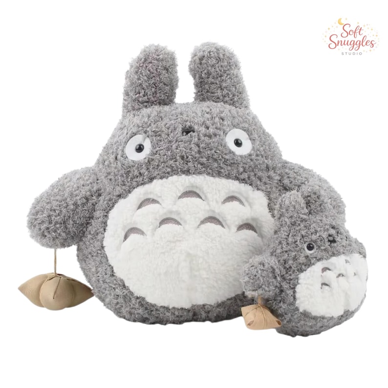 May include: A plush gray Totoro stuffed animal with a white belly and large eyes. A smaller Totoro plush is next to the larger one. The plush toys have a soft, textured appearance. The image includes the text "Soft Snuggles Studio".