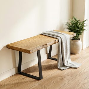 May include: A wooden bench with a natural edge and black metal legs. A gray blanket is draped over the bench. A potted plant sits to the right of the bench. The bench is made of light-colored wood.
