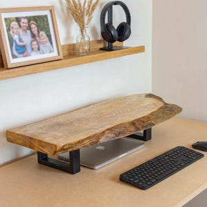 Può includere: Un supporto per monitor in legno con bordo naturale, caratterizzato da una venatura marrone chiaro e un profilo organico irregolare. La lastra di legno è sostenuta da due gambe rettangolari in metallo nero opaco, creando una base rialzata.