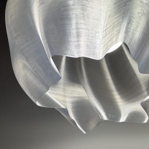 May include: A translucent, white pendant light shade with a wavy, organic, and rippled texture. The material appears thin and slightly flexible, featuring visible horizontal striations throughout the surface, with a bright light source visible inside.