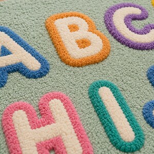 May include: A tufted rug featuring a light green background with raised, textured letters of the alphabet. The letters A, B, C, H, I, and J are visible, each outlined in vibrant colors including blue, orange, purple, pink, and teal with white centers.