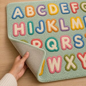 May include: A rectangular light green rug featuring a tufted alphabet pattern with letters in various colors including blue, orange, purple, yellow, pink, and green. The rug has a textured surface and a light-colored, non-slip backing material.