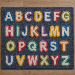 May include: A rectangular dark gray rug featuring the English alphabet in a tufted, textured style. Each letter is rendered in a bold, rounded font with a cream-colored center and a vibrant, multi-colored outline in shades of blue, orange, purple, yellow, red, and green.