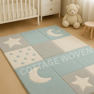 May include: A rectangular, patterned rug with a celestial theme. The rug features squares in light blue, gray, and white, with star, moon, and polka dot designs. The text "COTTAGE WOVEN" is visible on the rug.