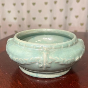 Può includere: Una ciotola in ceramica color verde menta chiaro, di forma rotonda, con un profilo basso e un'ampia apertura. La superficie esterna è decorata con un motivo in rilievo a forma di foglia che circonda il centro, con un bordo perlato.
