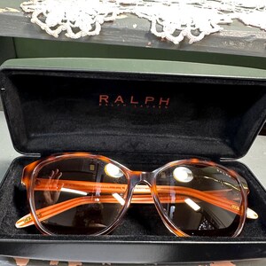 Puede incluir: Estuche de gafas de sol negro abierto con gafas de sol Ralph Lauren. Las gafas de sol tienen una montura con estampado de carey y lentes marrones. El nombre de la marca "RALPH" est&aacute; impreso en rojo en el estuche.