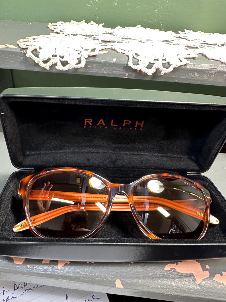 Puede incluir: Estuche de gafas de sol negro abierto con gafas de sol Ralph Lauren. Las gafas de sol tienen una montura con estampado de carey y lentes marrones. El nombre de la marca "RALPH" est&aacute; impreso en rojo en el estuche.
