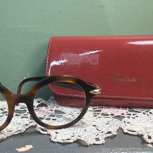 May include: A pair of round, tortoiseshell eyeglasses with a red, glossy eyeglass case. The case has gold lettering and a clasp. The glasses and case rest on a white lace doily.