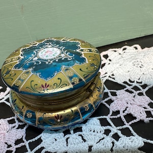 Op de afbeelding: Een vintage, blauwe en gouden glazen sieradendoos met een bloemenmotief en een scharnierend deksel. De doos is versierd met ingewikkelde details en heeft een delicate, vrouwelijke uitstraling.