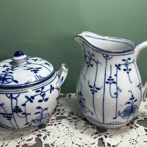 May include: A white and blue porcelain sugar bowl with a lid and a matching creamer. Both pieces feature a blue floral pattern and gold trim. The sugar bowl has two handles and a blue knob on the lid. The creamer has a handle and a spout.