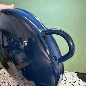 May include: A deep blue ceramic pitcher with a curved handle. The pitcher has a glossy finish and a rounded shape. The handle is a loop design. The pitcher is sitting on a white lace tablecloth.