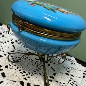 Puede incluir: Una caja de vidrio azul vintage con un diseño floral y una tapa con bisagras de metal dorado. La caja se asienta sobre un soporte de metal dorado con tres patas.