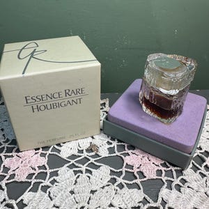 Op de afbeelding: Een vintage parfumflesje met een getextureerd glazen ontwerp, gevuld met een donkere vloeistof, staat op een paars kussen in een grijze doos. Daarnaast staat een beige doos met de tekst "Essence Rare Houbigant" en "The Perfume .25 FL OZ".