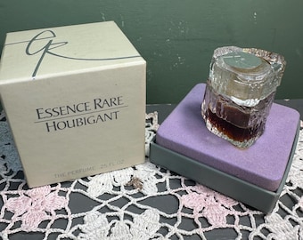 Può includere: Un flacone di profumo vintage con un design in vetro strutturato, riempito con un liquido scuro, è posto su un cuscino viola all'interno di una scatola grigia. Accanto c'è una scatola beige con il testo "Essence Rare Houbigant" e "The Perfume .25 FL OZ".