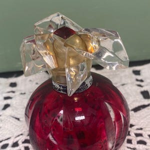 May include: A round, red glass perfume bottle with a faceted crystal stopper. The bottle has a gold-colored neck and a dark red, textured body. The stopper is clear with a red accent. The bottle is sitting on a white lace surface.