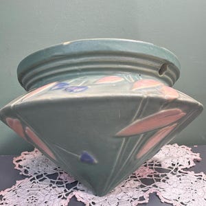 May include: A teal-colored ceramic planter with a geometric design. The planter features pink and blue floral accents. The top rim has a ribbed texture. The planter is likely for indoor use and adds a touch of vintage charm to any space.