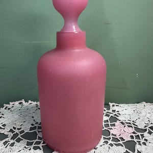 May include: A vintage pink glass perfume bottle with a bulbous body and a rounded stopper. The bottle has a frosted finish, and the stopper is a lighter shade of pink. The bottle is displayed on a white lace doily.