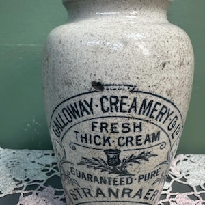 Może przedstawiać: Antyczny kamienny słoik na śmietanę z nakrapianym, kremowym wykończeniem. Słoik posiada czarną etykietę z napisem "Galloway Creamery Co. Fresh Thick Cream, Guaranteed Pure, Stranraer". Słoik ma lekko rozszerzoną krawędź.