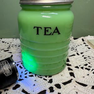 Könnte beinhalten: Eine vintage Teedose aus grünem Glas mit einem silbernen Deckel. Die Dose hat ein geripptes Design und das Wort "TEA" ist auf der Vorderseite aufgedruckt.