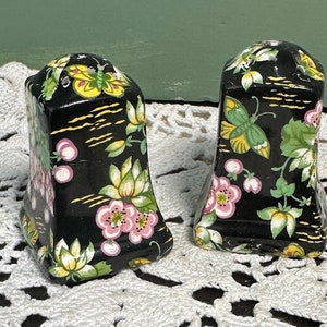 May include: Two black ceramic salt and pepper shakers with floral and butterfly designs. The shakers feature pink flowers, yellow butterflies, and green leaves. The background is black with gold accents.