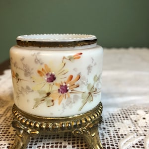 Może przedstawiać: Biały porcelanowy słoik z pozłacanym brzegiem i kwiatowym wzorem, stojący na złotej metalowej podstawce z trzema nóżkami.