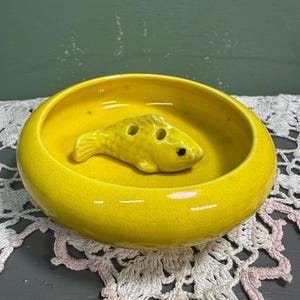 May include: A bright yellow ceramic ashtray with a fish-shaped design in the center. The ashtray has a round shape with a slightly raised edge. The fish has two holes and a black eye. The ashtray is sitting on a white and pink lace doily.