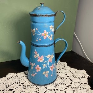 May include: A vintage, pale blue enamel coffee pot with a floral design. The pot features pink and white flowers with green leaves. It has a lid, a spout, and a handle. The coffee pot is sitting on a white lace doily.