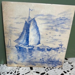 Può includere: Una piastrella di ceramica blu e bianca con un'immagine disegnata a mano di una barca a vela su uno specchio d'acqua con un mulino a vento sullo sfondo.