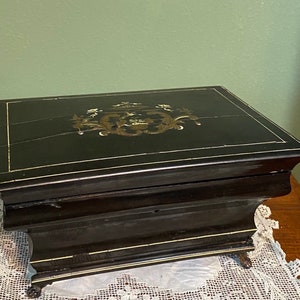 May include: A black antique box with gold trim and a decorative gold emblem on the lid. The box has a rectangular shape and sits on small, ornate feet. The box is likely used for storing jewelry or other small items.