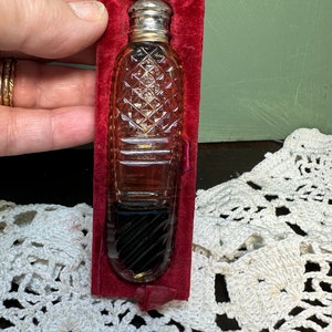 May include: A vintage perfume bottle with a silver cap and a clear, diamond-cut glass body. The bottle contains a dark liquid and is presented in a red velvet holder. A small red ribbon is attached to the holder.