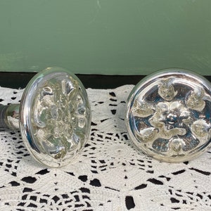 May include: Two silver bottle stoppers with a floral design. One stopper is oval-shaped, and the other is round. The stoppers have a reflective, mirrored finish and a dark gray rubber seal. The background is a white lace doily.