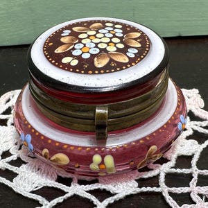 Puede incluir: Un pequeño tarro de vidrio rosa redondo con una tapa blanca y un diseño floral. La tapa está decorada con un patrón floral marrón y blanco. El tarro tiene una bisagra de latón.
