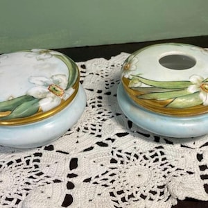 May include: Two oval-shaped, light blue ceramic containers with lids. The lids and rims are adorned with gold trim and painted floral designs featuring white flowers and green leaves. One container has a hole in the center.