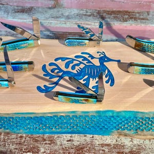 May include: A wooden display featuring several pocket knives with blue and silver accents. The knives are arranged around a blue seahorse design. The display has a turquoise border and a black box with white text.