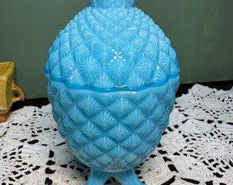 May include: A light blue glass pineapple-shaped candy dish with a scalloped top and three feet. The dish is sitting on a white crocheted doily.