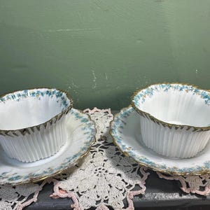 May include: Two white teacups and saucers with a blue floral pattern and gold trim. The teacups have a fluted design and sit on matching saucers. The set is displayed on a lace doily, suggesting a vintage or antique aesthetic.