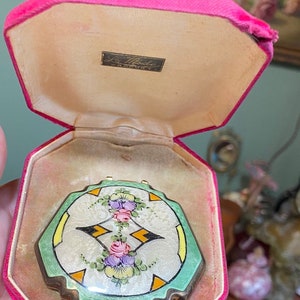 May include: A vintage green and gold compact with a floral design and the text "La Mode" in the center. The compact is in a pink velvet case.