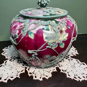 May include: A vintage ceramic jar with a lid, featuring a floral design with pink roses and green leaves. The jar has a white and silver trim and a green base.