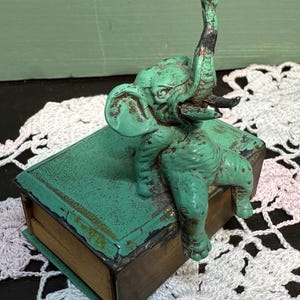 以下が含まれることがあります： 緑色のヴィンテージマッチボックスに、金属製の象の置物が置かれています。象は後ろ足で立ち、鼻を上げています。マッチボックスは木製の底になっています。