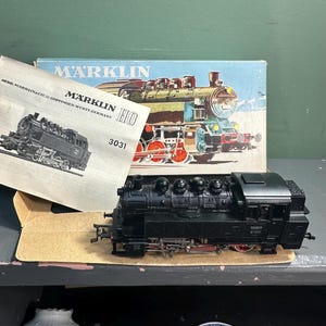 May include: A black Marklin HO model train with red wheels sits in front of its box and instruction manual. The box features a colorful illustration of a train. The instruction manual has a black and white drawing of a train and the text "Marklin HO 3031".